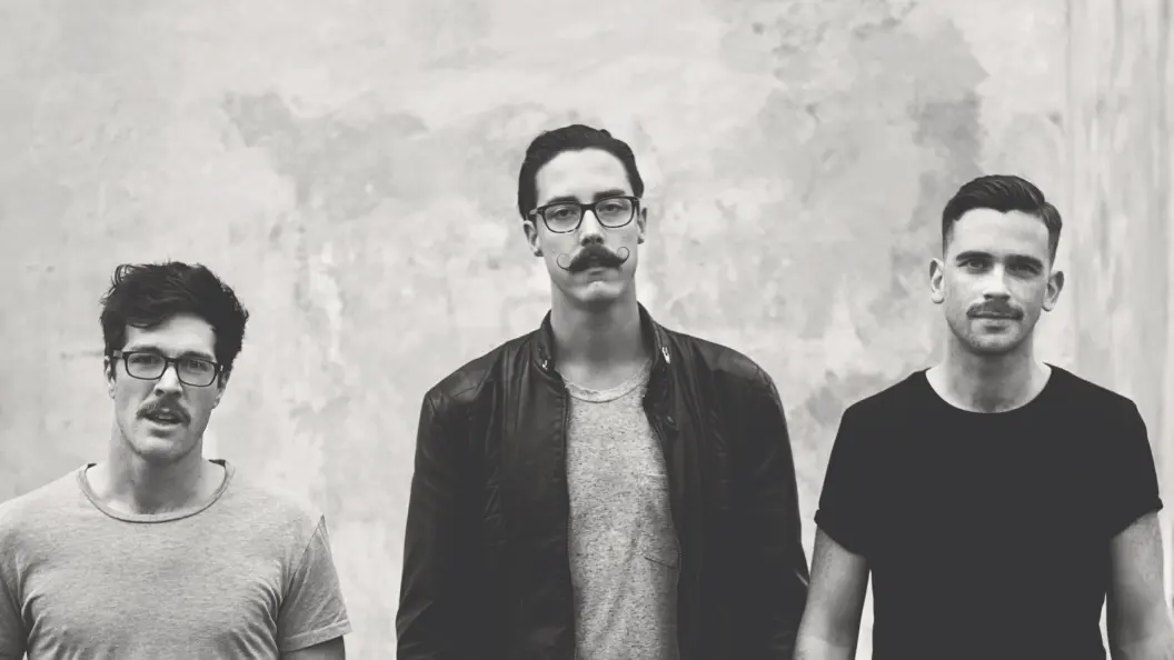 Portrait of three men with moustaches against concrete wall.
